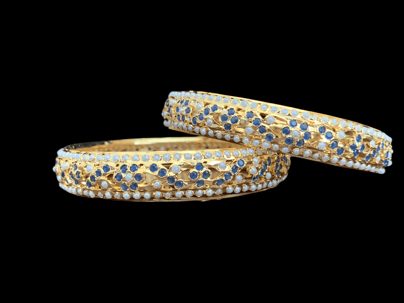 Two of Deccan Jewelry’s leela bangles, intricately detailed in gold with patterns of pearls and sapphire-blue stones. The two bangles are stacked atop one another.