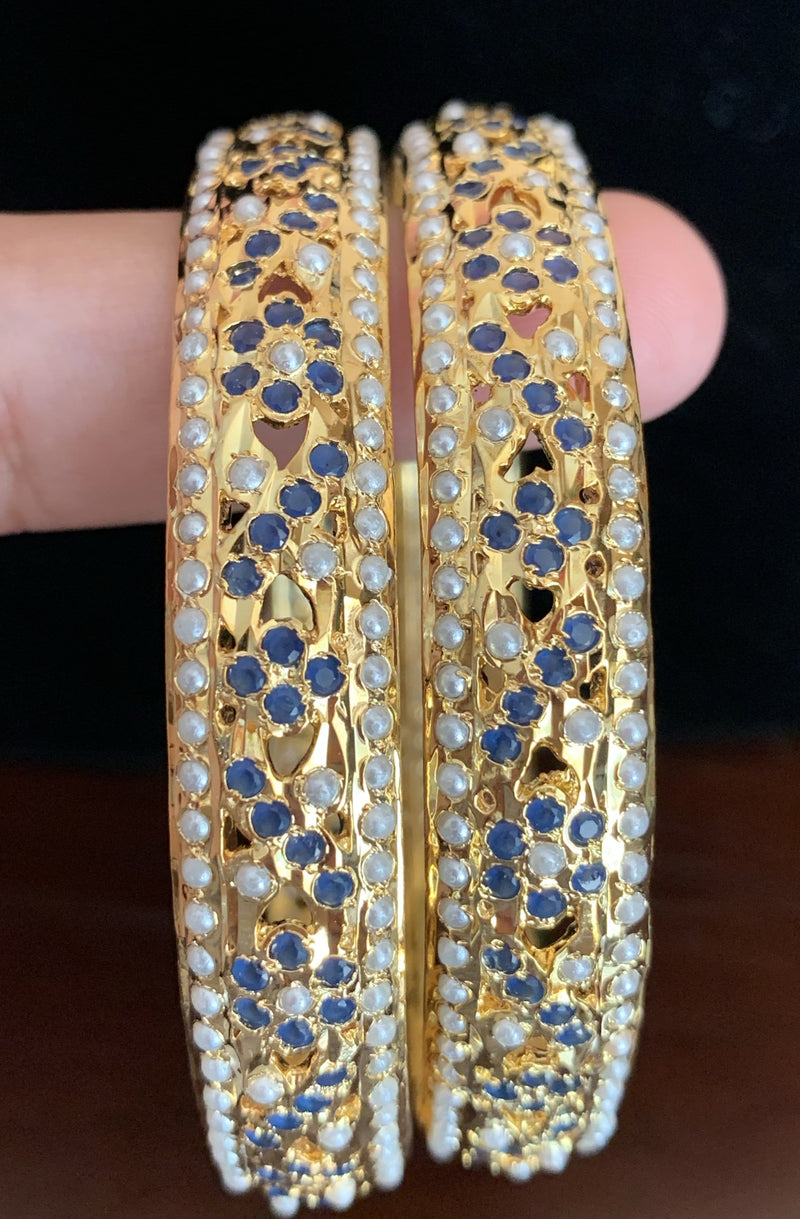 Two of Deccan Jewelry’s leela bangles, intricately detailed in gold with patterns of pearls and sapphire-blue stones. Both bangles are hung around a finger.